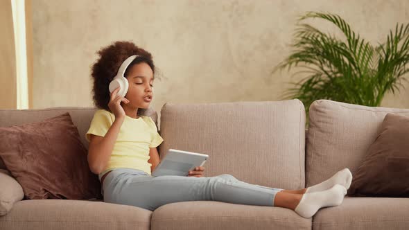 Portrait Little African American Girl in Big White Headphones Listens Music Using Digital Tablet alt