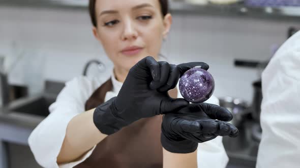 Female Pastry Chef in Black Gloves Holds a Handmade Chocolate Sphere in Her Hands alt