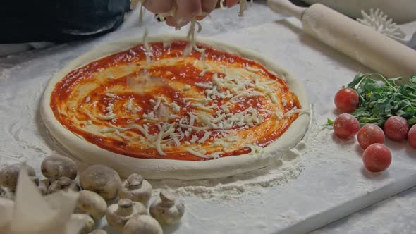 Professional Chef Pours Grated Cheese Onto a Pizza Base with Tomato Paste on the Table Decorated alt