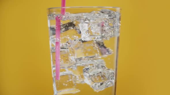 Somebody Mix Water Drink with Drinking Straw. Glass Full of Fizzy with Ice Cubes Yellow Background alt