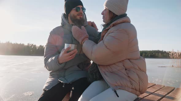 Cute Couple Drink Hot Tea From Thermos and Kiss Sitting on a Dock in Winter
