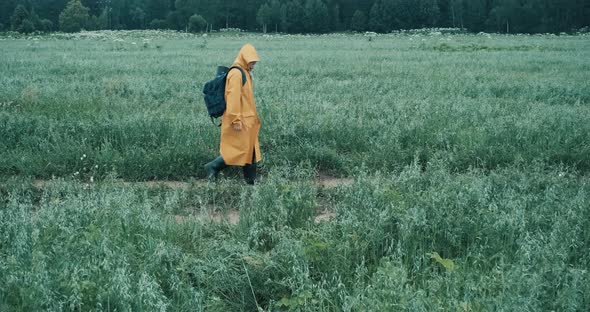 Man in a Yellow Raincoat and a Backpack Walks Through a Field with Grass