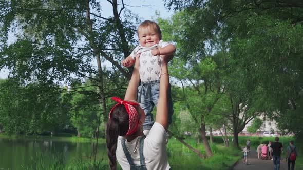 Happy Cheerful Young Mother Holding His Small Lovely Son High in Hands and Laughing in the Park alt