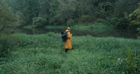 Hiker in a Yellow Raincoat with a Backpack in the Tall Grass Next to the River