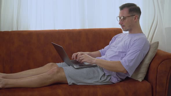 A Man with Glasses Works at a Laptop Sitting on the Couch From Home Remote Work and Study