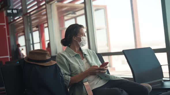 A Young Woman in a Facial Mask Sitting at the Airport and Using Her Smartphone alt