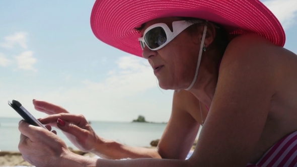 Middle Aged Woman In Hat And Sunglasses Uses Mobile Phone On a Beach