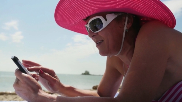 Middle Aged Woman In Hat And Sunglasses Uses Mobile Phone On a Beach