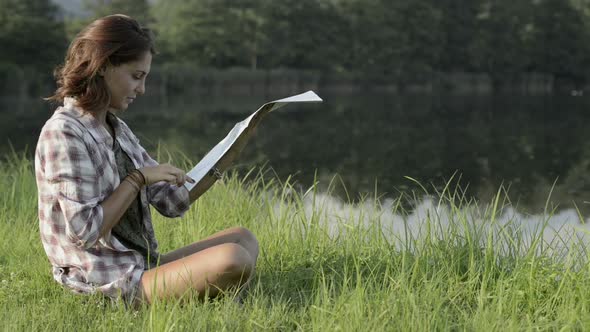 Young Woman in Nature Seated on Grass Look at Map on Lake Shore in Sunny Summer Day Outdoor