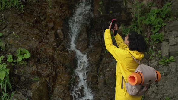 Woman Tourist is Filming Small Waterfall in Forest By Camera of Smartphone alt