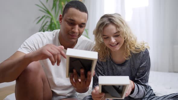 Happy Young Couple Talking Smiling Looking at Pictures Sitting in Bedroom in the Morning alt