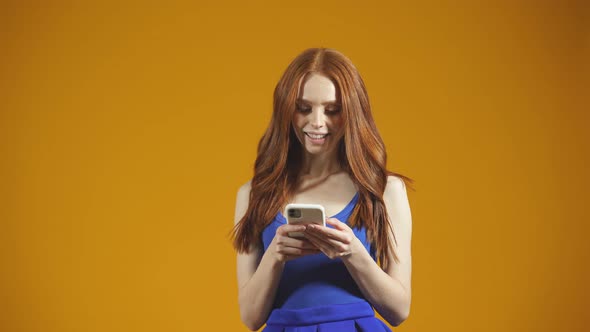 Young Woman with Long Red Hair and a Smartphone in Her Hands Stands on an Isolated Background in the