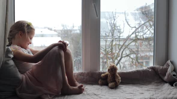 Girl Sitting on a Windowsill with a Teddy Bear and Smiling