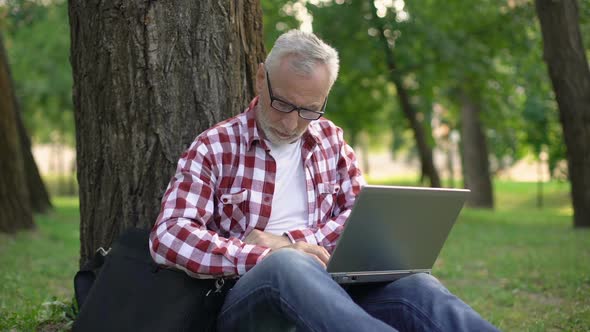 Senior Man Sitting on Grass and Booking Tickets Online, Male Resting in Park alt