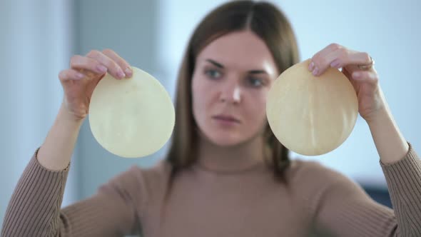 Rack Focus From Breast Implants in Female Hands to Thoughtful Caucasian Young Woman Choosing alt