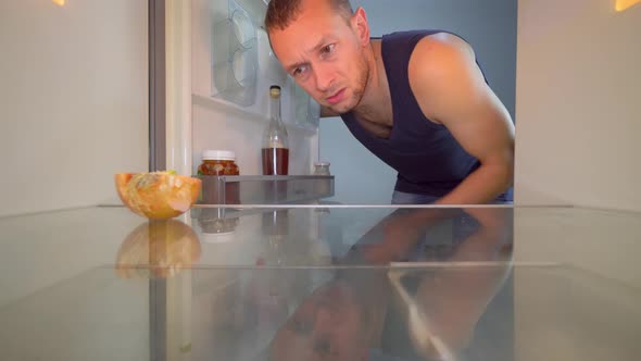 man looks into refrigerator, looks at onion and takes a bottle of alcohol. alt
