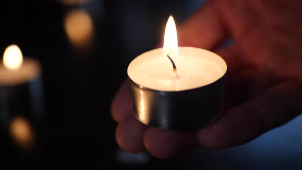 A hand holding a small tealight candle with the flames of many burning in the background during a ca alt