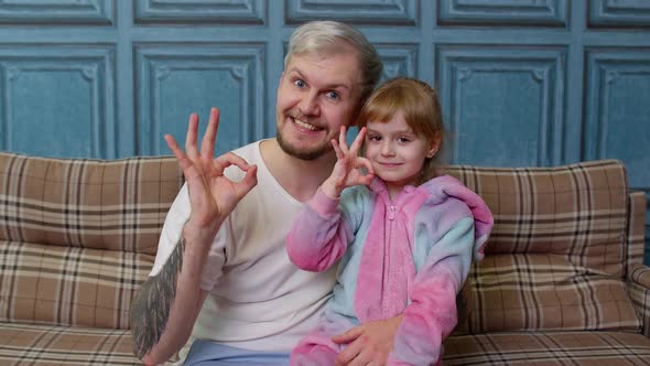 Father and Little Child Daughter Kid in Pajamas Sit on Couch in Room Smiling Showing Ok Gesture Sign alt