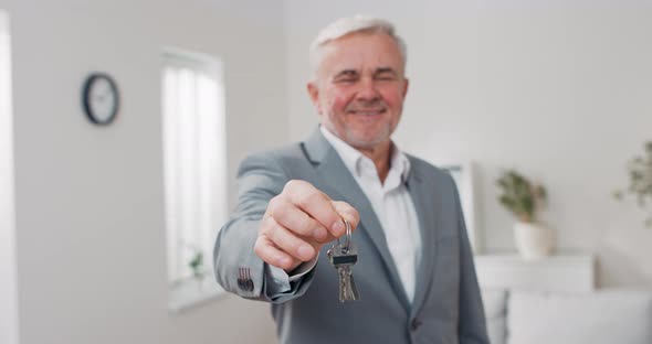 Shot of Apartment Keys Extended on Palm of Hand Toward Camera Grayhaired Mature Man Dressed in Suit alt