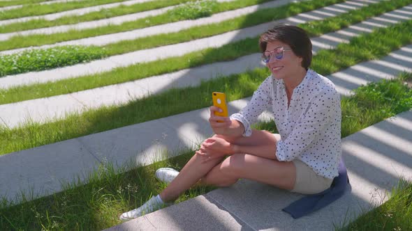 Woman with Colorful Sunglasses Sits on Lawn in Urban Park and Makes Selfie on Smartphone alt