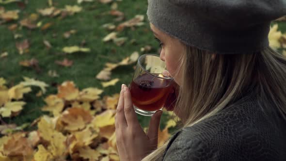 Woman Sits on Autumn Maple Leaves and Drinks Hot Mulled Wine with Lemon in Park Outdoors
