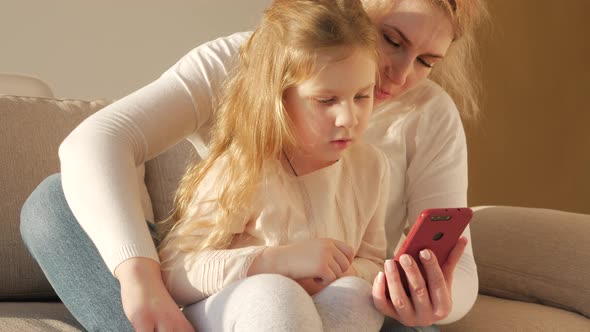 Young mom and her little daughter are playing a smartphone and smiling while sitting on sofa at home