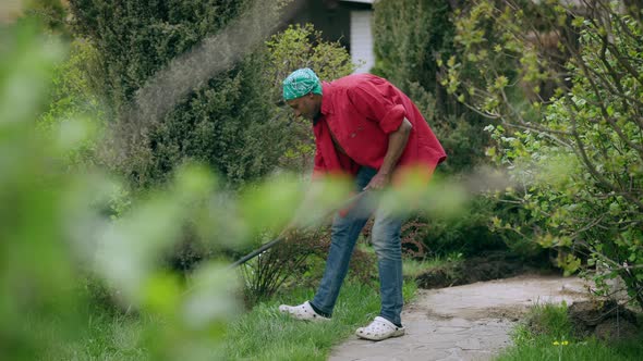 Young African American Man in Jeans and Red Shirt Raking Garden on Hot Summer Day Outdoors and alt