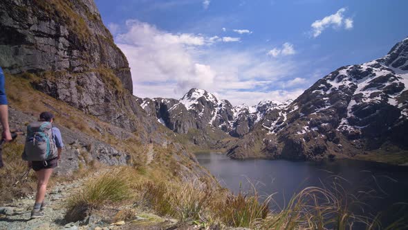 Static, hikers walk above alpine lake through snow capped mountain landscape, Routeburn Track New Ze alt