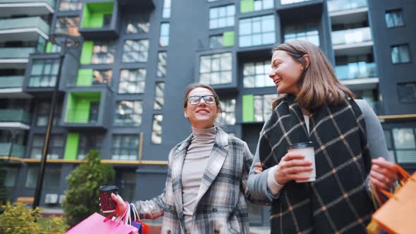 Two Happy Women Walk with Shopping Bags and Takeaway Coffee After a Successful Shopping and Talk alt
