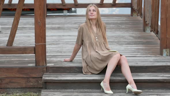 Young attractive blonde girl in a dress sits on a wooden pier