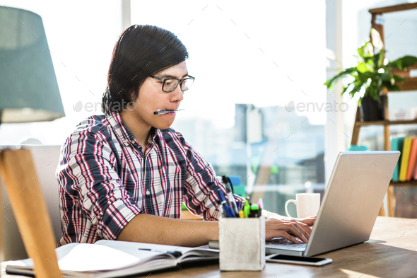Hipster businessman with pen in mouth using laptop in office