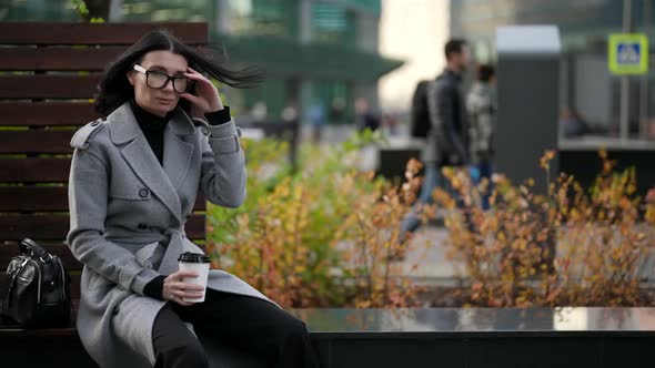 Elegant Lady is Sitting on Bench in Park and Drinking Coffee From Cardboard Cup alt