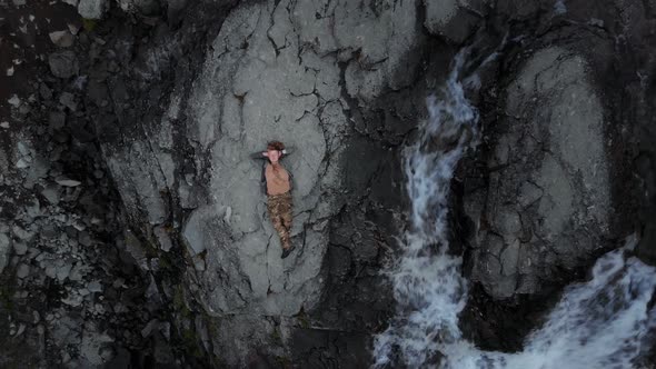 Man Lies with Hands Under His Head on Giant Boulder with Mountain River Around