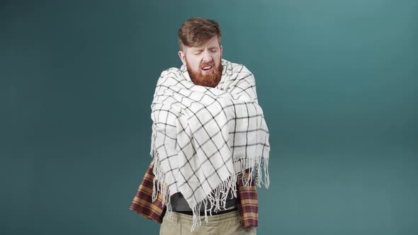 A Bearded Young Man in White Scarf Stands Crossing His Arms and Sneezes alt