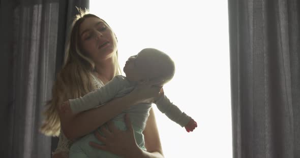 Happy Mother Swings Her Newborn Son and Caresses Him in Room at Bright Window alt
