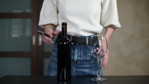 The girl comes to the table with a bottle of wine and a glass and starts to open wine alt