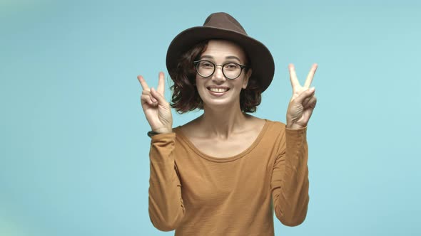 Funny Hipster Girl in Glasses and Hat Showing Peace Victory Signs and Smiling at Camera Standing