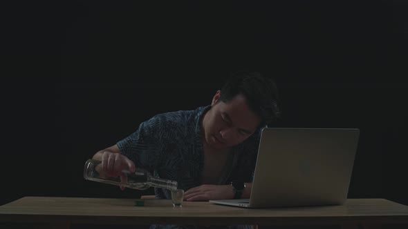 Asian Man Drinking Vodka While Working On A Laptop In Black Background alt