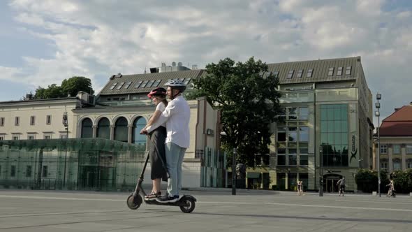 Young Man Rides His Girlfriend on an Electric Scooter in the City Square