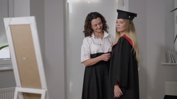Proud Caucasian Mother and Daughter in Graduation Outfit Admiring Reflection in Mirror Standing in alt