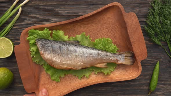 Hands are Placing a Wooden Plate with a Grilled Red Char Fish on the Black Table alt