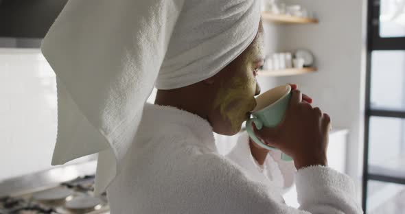 Happy african american woman with beauty mask on face, drinking coffee in kitchen alt