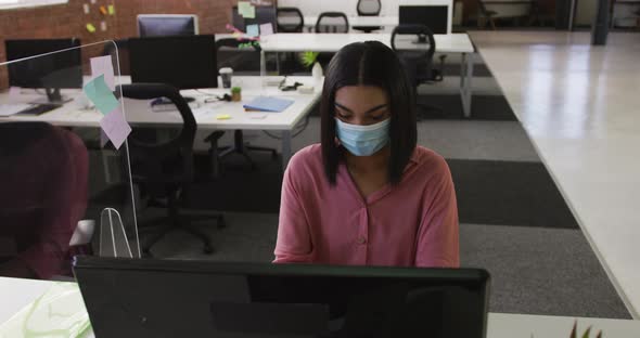 Mixed race businesswoman wearing face mask sitting at desk using computer alt