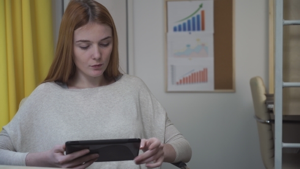 Woman Sits At The Workplace, In The Hands Holding The Tablet. alt