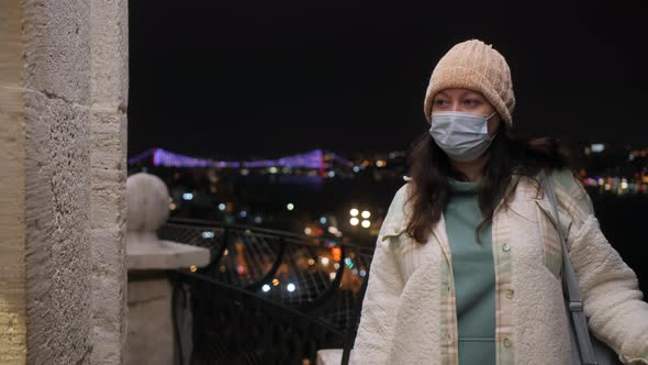 Woman in Mask Leaning at Railing with Istanbul Cityscape Behind at Night alt