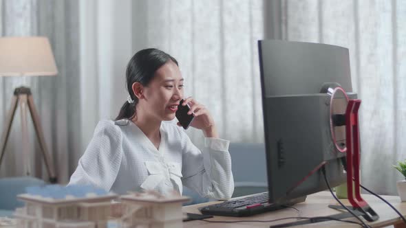 Asian Woman Engineer With The House Model Talking On Smartphone While Working On A Desktop At Home alt