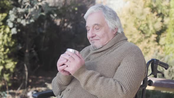 Side View Portrait of Elderly Caucasian Man Drinking Hot Tea As Sitting in Wheelchair Outdoors. Old alt
