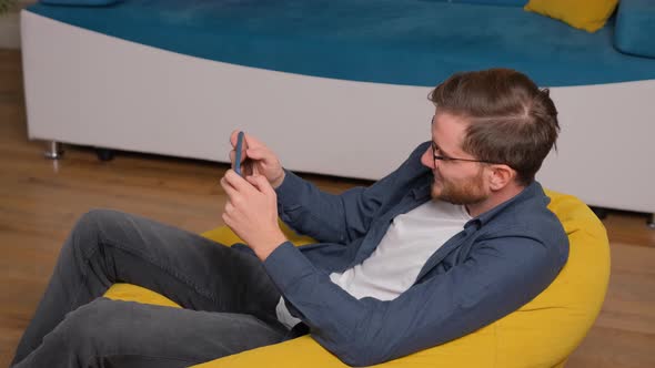 Young Man Playing Games on Phone, Sitting on a Bean Bag at Home. alt