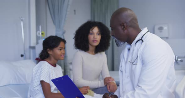 African american senior male doctor with clipboard talking to mother and daughter at hospital alt