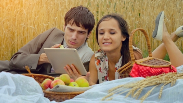  Happy Couple In Love Sitting On Picnic In Wheat Field Use Tablet And Eating Apples. 